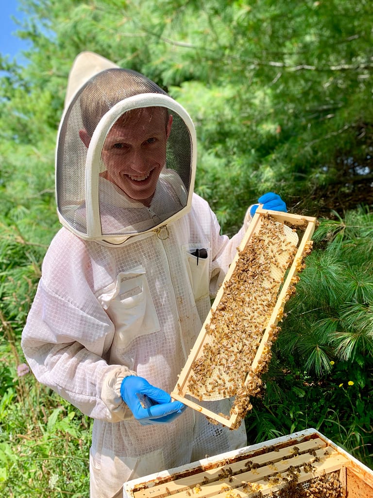 Ben DeMoras in a bee suit showing a frame of bees from an open hive.