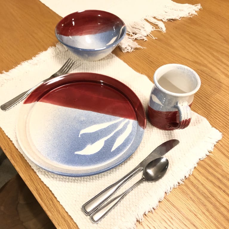 A place setting showing a plate, mug, and bowl, each in firebrick red and blue glaze